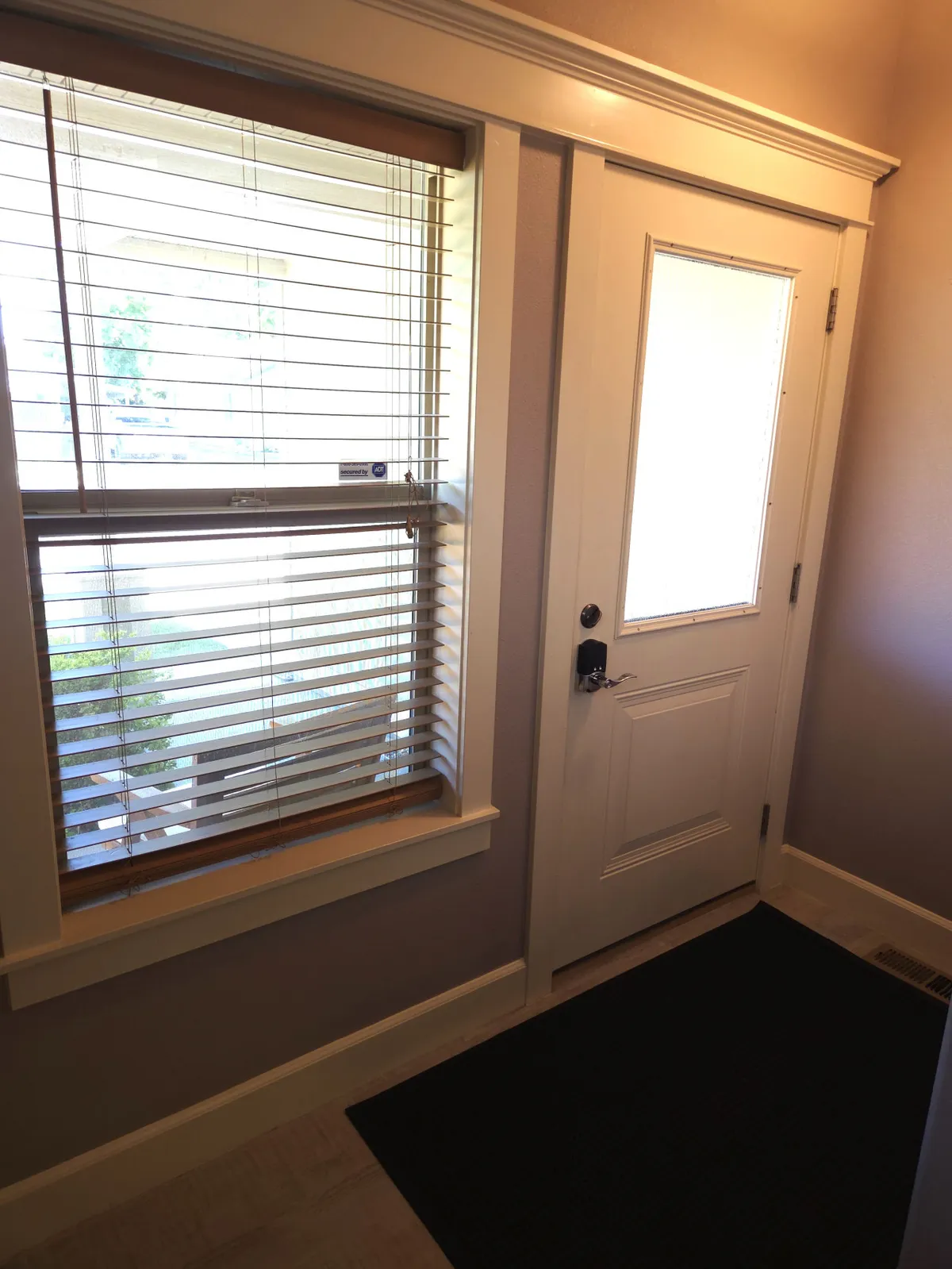 This image shows the front door of the home, flanked by a window with blinds and an entry mat. The entryway is bright and welcoming, setting a positive first impression for guests and residents alike. The scene suggests a warm, family-friendly atmosphere where people come and go, emphasizing connection to the outside world.