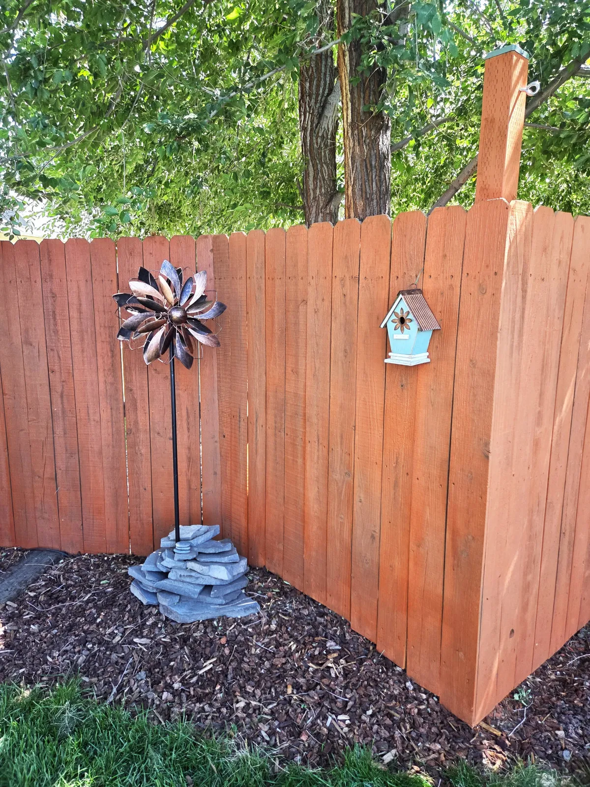 A playful and serene corner of a backyard with a decorative windmill, a charming birdhouse, and a wooden fence enclosing the space. The garden area is lush and sunny, showcasing features that invite gardening, birdwatching, and relaxed outdoor moments.