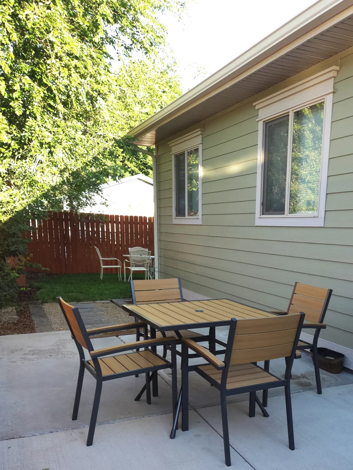 A wooden patio table with metal chairs sits amid lush greenery and a striking red wooden fence. The area is dappled with sunlight, creating a vibrant, cozy setting for al fresco meals and casual gatherings.