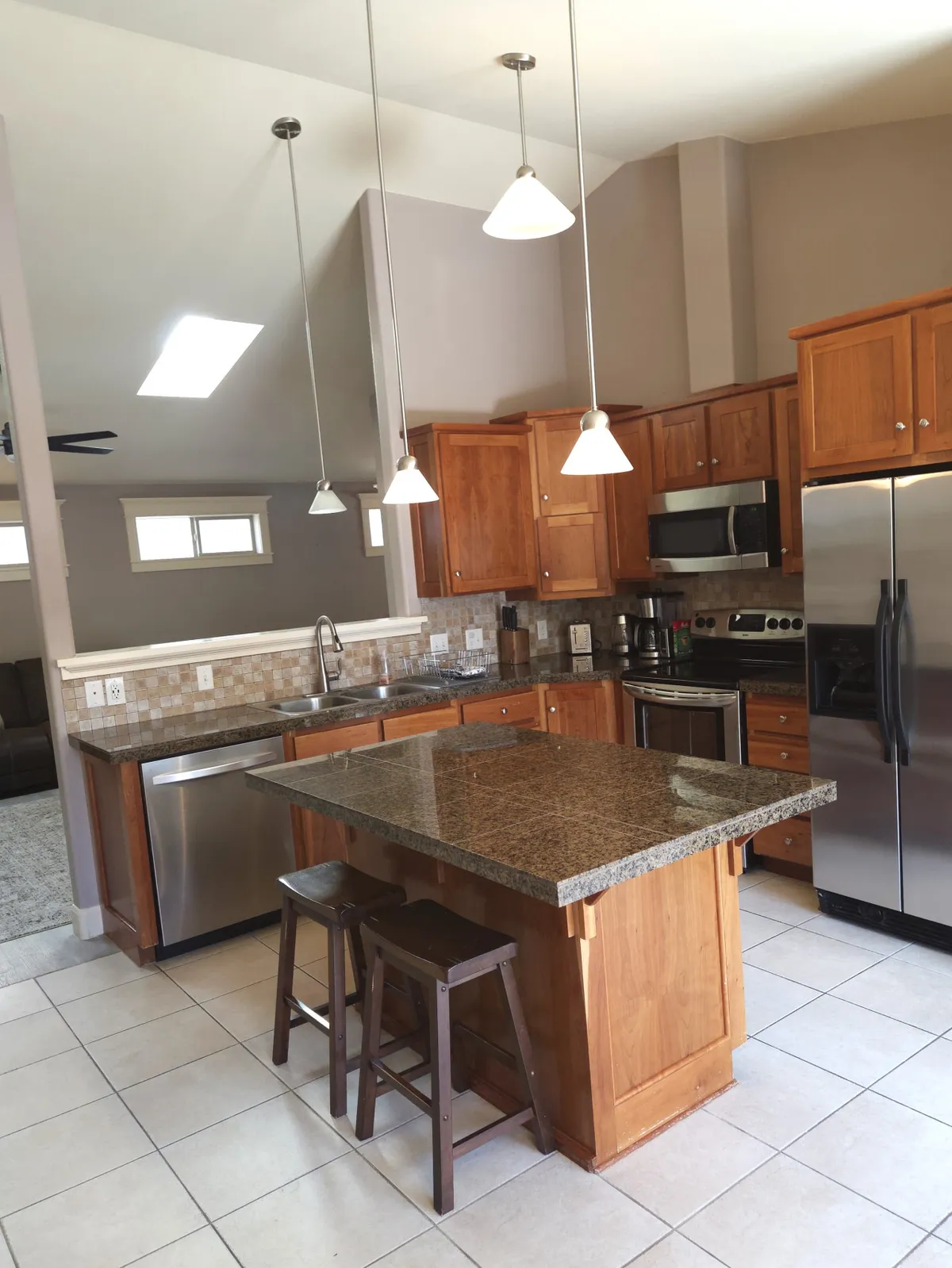 A spacious kitchen featuring a granite island countertop, wooden cabinets, and stainless steel appliances, illuminated by stylish hanging light fixtures. The setting is warm and welcoming, clearly designed for both cooking and socializing, with enough space for family gatherings and casual dining.