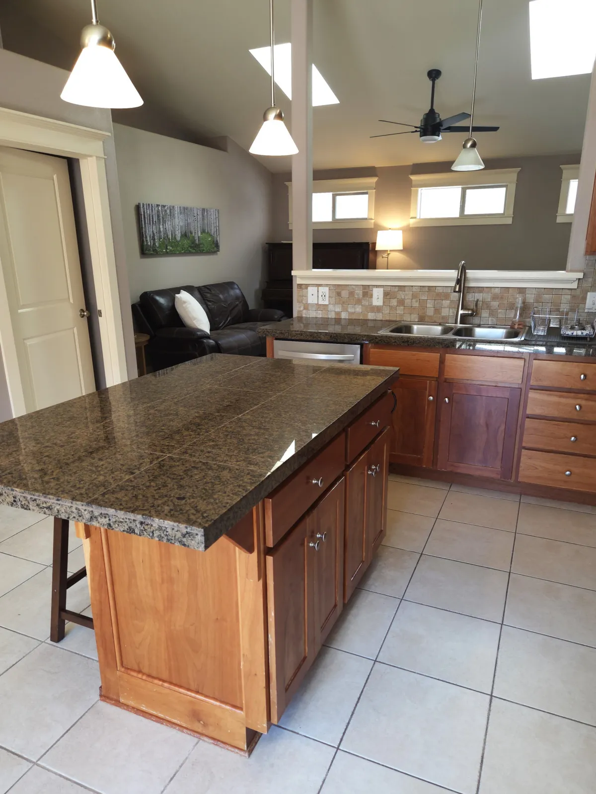 This inviting interior showcases a sunlit open-concept kitchen with a gleaming granite island, warm wood cabinetry, and a cozy living area complete with a plush couch and cheerful artwork. The space is clearly designed for gathering, cooking, and creating memorable moments with family or friends.