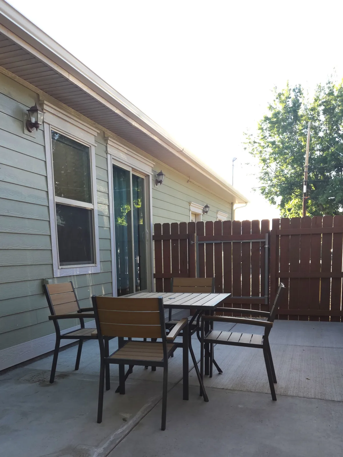 A cozy outdoor patio setup featuring a table and chairs just outside the home, framed by a wooden fence and nearby windows. The space is ideal for sipping morning coffee, sharing meals, or unwinding with loved ones while enjoying the fresh air.