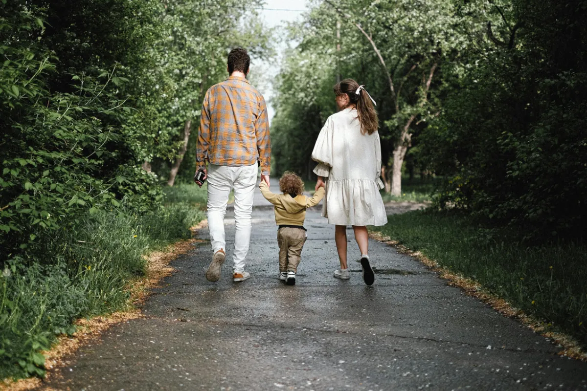 A cheerful scene of a family walking together along a tree-lined path, surrounded by lush greenery and dappled sunlight. The overhead canopy creates a pleasant, inviting atmosphere for outdoor exploration and family bonding.