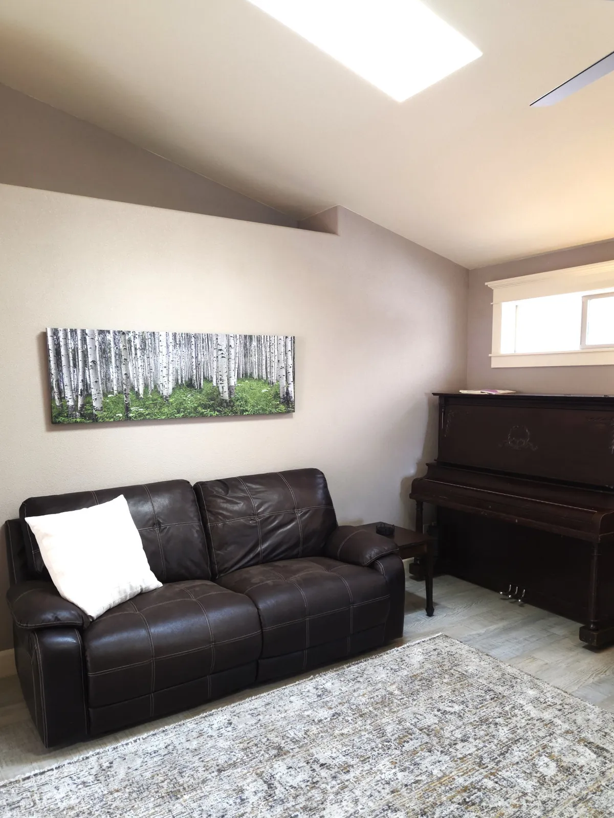 A living room featuring a brown leather sofa and a piano, with birch tree artwork above the piano. The space is arranged for relaxation and socializing, with comfortable seating and an artistic touch.