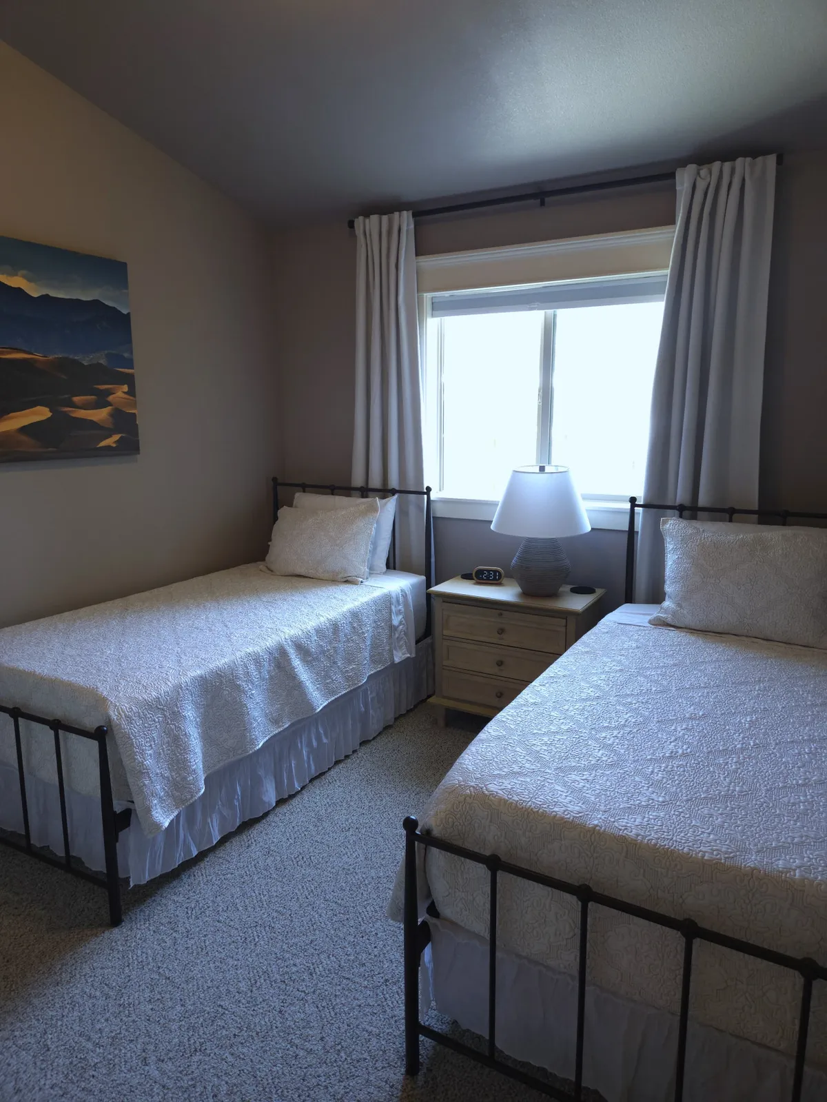 A comfortable bedroom with twin beds, a window draped with curtains, a nightstand with a lamp and clock, and a painting of mountains on the wall. The room feels inviting, perfect for families or groups needing personal space.