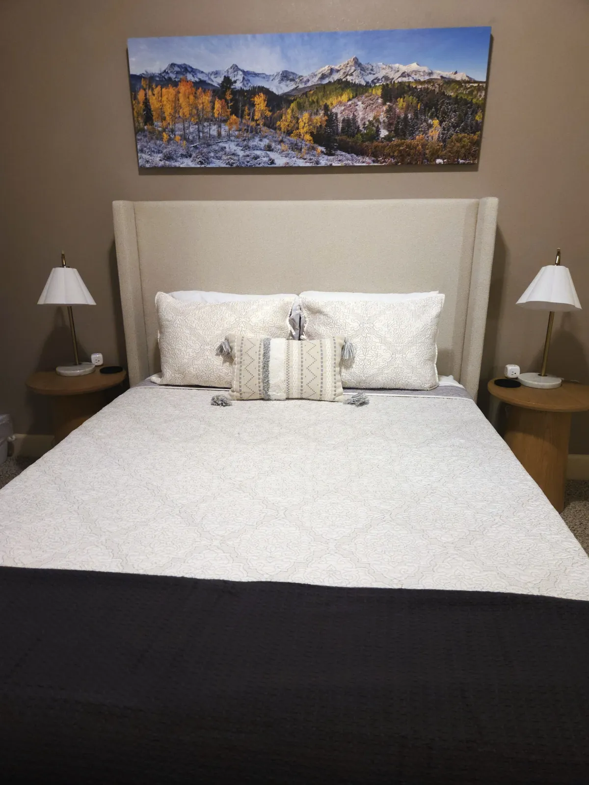 A calm bedroom featuring a bed with decorative pillows, mountain landscape artwork above, and lamps on nightstands flanking the bed. The atmosphere is peaceful and soothing, offering a sense of escape and comfort.
