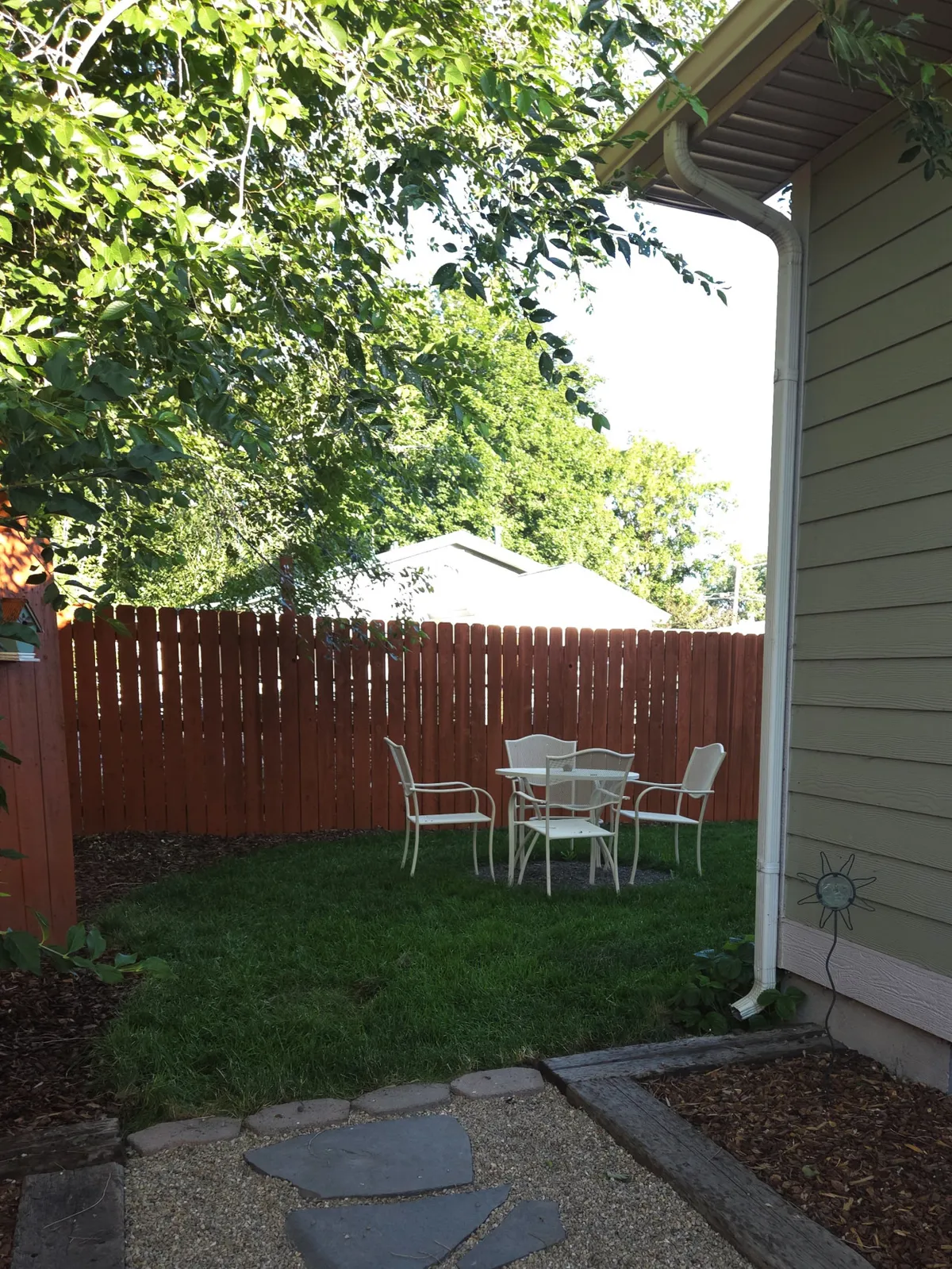 A tranquil backyard scene featuring a white outdoor dining set placed on a lush green lawn, bordered by a sturdy wooden fence. The space is bathed in natural sunlight, inviting guests to gather, dine, and relax outdoors. The open fenced yard hints at safety for children and pets, while the setup suggests family meals or evening gatherings under the stars.