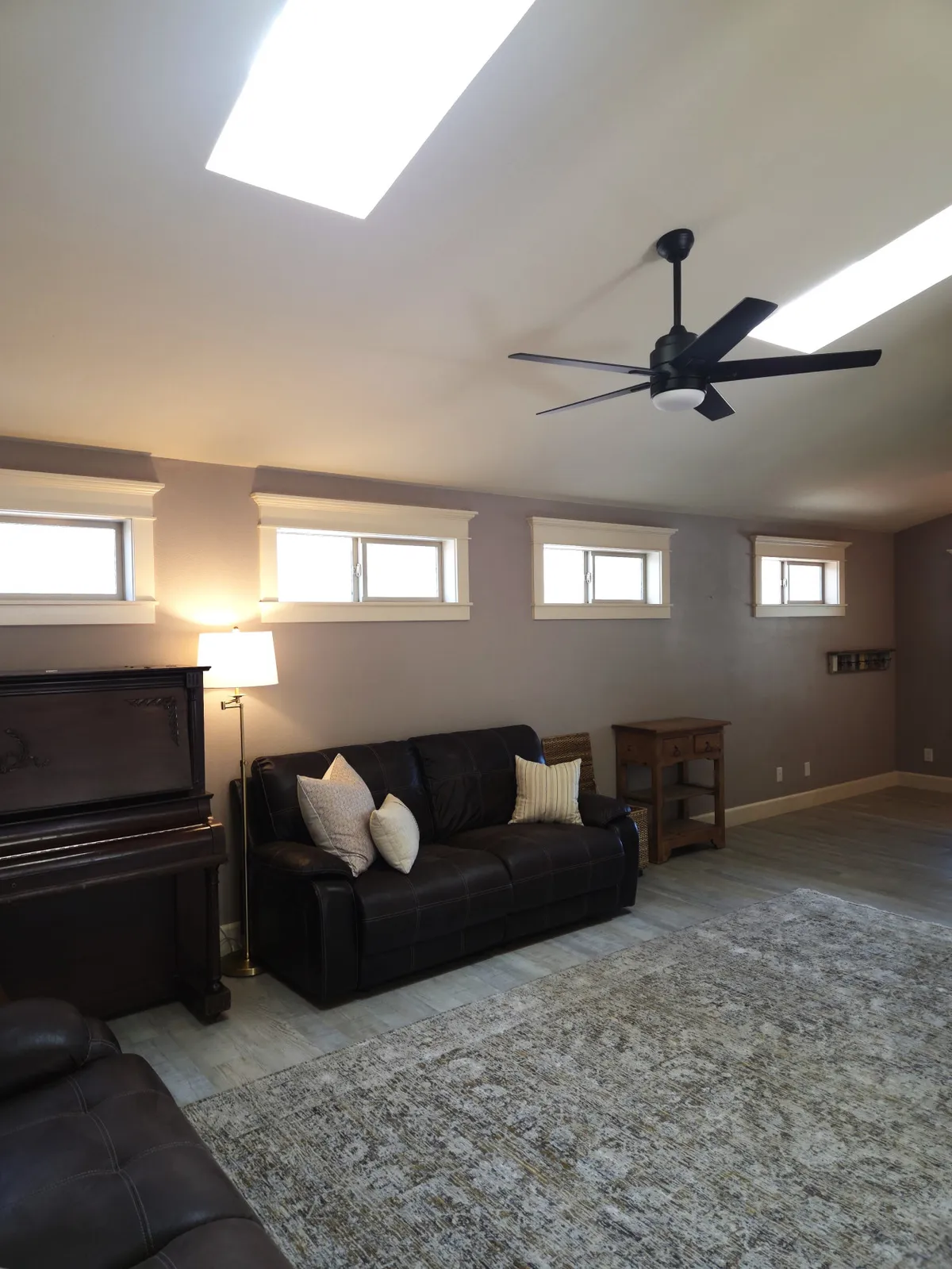 A cozy, well-lit living room with a sofa, piano, table, stylish light fixture, and a soft rug, creating a space ideal for relaxing, playing music, or gathering for game night.